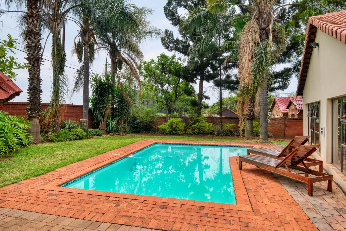 Brick paver pool deck with two wooden sun loungers beside a stucco home with a tile roof