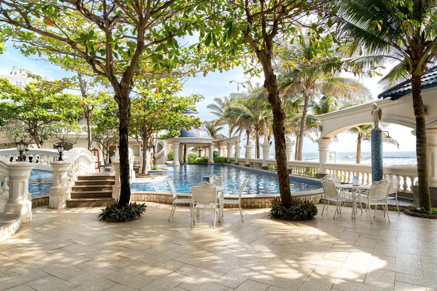 Clean stone-paver pool deck with white patio tables, palm trees and the ocean beyond